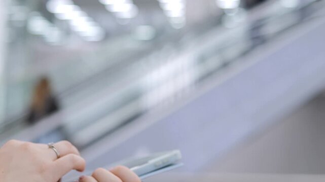 Hand holds phone while riding escalator. Blurred background shows indoor transit space. Person uses device during movement. No animals or plants visible