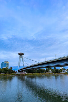 Most SNP bridge over Danube River in Bratislava. Its observation tower and cable stays rise beside modern buildings under a pale evening sky.