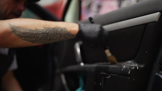 Close-up view of a tattooed male worker cleaning the interior door panel of a car with a specialized brush and foaming cleaner, meticulously restoring the vehicle's interior