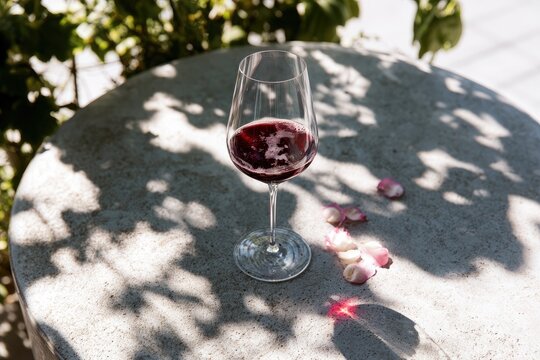 A glass of red wine sits on a textured surface with scattered flower petals and dappled sunlight