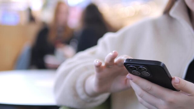 Close-up of hands scrolling on a smartphone in a cafe. Person wears light-colored sweater, focused on screen. Background shows blurred cafe patrons. Ideal for social media, digital marketing
