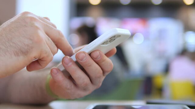 Close-up hands tap phone screen. Focus on fingers scrolling or texting. Background blurred with people and colorful surroundings. Suggests modern communication or multitasking in public