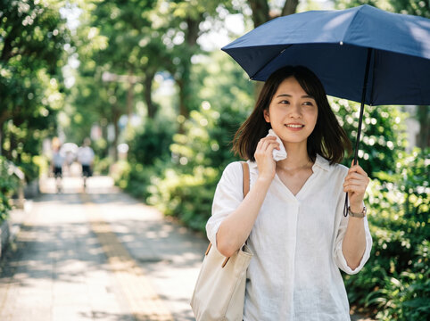 木漏れ日の並木道で青い日傘を差し、タオルで汗を拭いながら微笑む女性