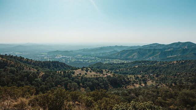 Landscape view of hills and valleys with distant mountains, clear sky, and a mix of greenery and dry terrain.