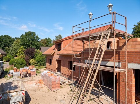 Modern two-story red brick house under construction with scaffolding, yellow ladder, and temporary fencing on sunny day.