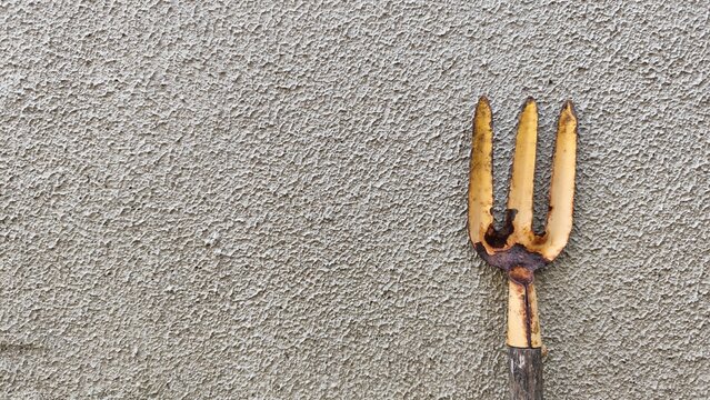A close-up shot of an old, used hand garden fork with worn metal tines placed against a textured grey concrete wall. Ideal for gardening themes, agriculture concepts, DIY home tools, backyard farming,