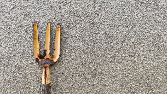A close-up shot of an old, used hand garden fork with worn metal tines placed against a textured grey concrete wall. Ideal for gardening themes, agriculture concepts, DIY home tools, backyard farming,