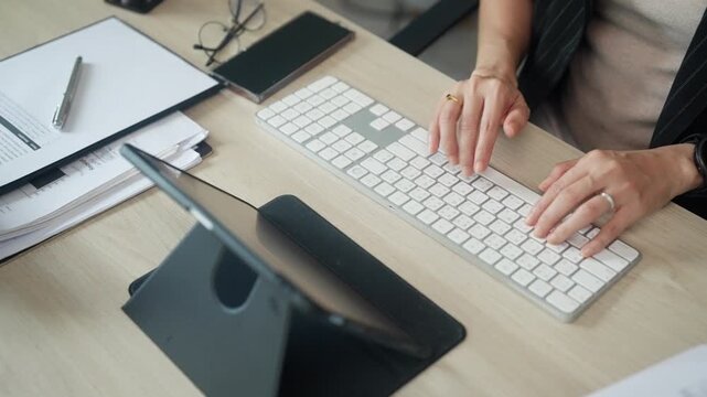 Overhead view of office employee typing on a wireless keyboard with documents and digital devices on a clean workspace. Suitable for administration, office management, business operations, and profess