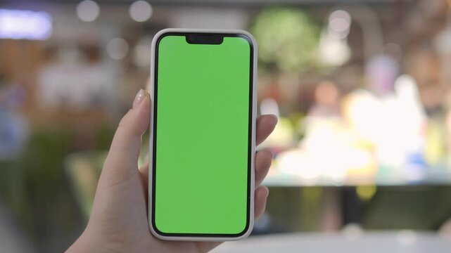 Hand holds smartphone with green screen. Background blurred, people moving casually. Ideal for app demos, digital content, or mockups. Focus on phone, not surroundings