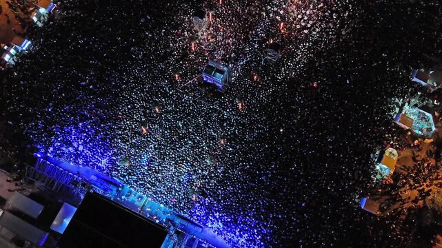 Aerial around view of crowd at night music concert. People gathered at festival outdoor stage with lights and screens, 4k