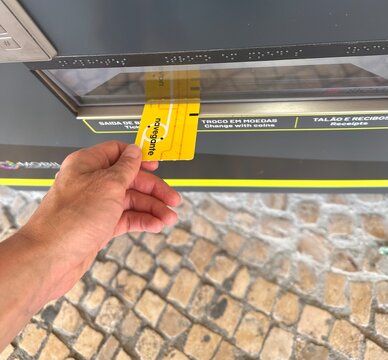 Passenger collecting a yellow Navegante travel ticket from an automated railway vending machine with Braille labels above traditional Portuguese stone pavement, 5 April 2026, Porto, Portugal
