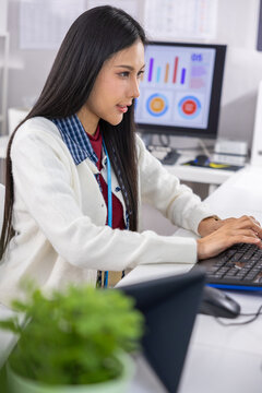 Asian woman office worker computer keyboard, focused employee typing at desk with chart monitor, indoor workplace with plant and business data screen Vertical