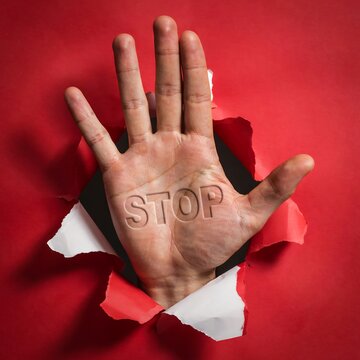 Human hand with the word stop written on the palm breaking through a torn red paper surface as a sign of warning or prohibition