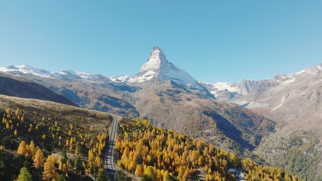 Aerial drone view of a scenic railway line curving through a colorful autumn forest with Matterhorn peak in Zermatt, Switzerland. Bright sunny day alpine landscape.