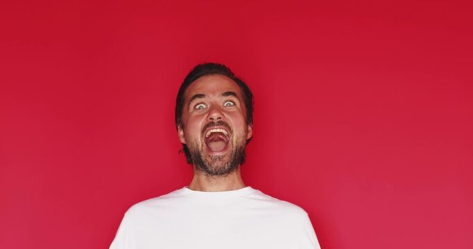 Handsome bearded man wearing white t shirt smiling happily before widening eyes and shouting with extreme shock intense surprise facial expression isolated on red studio background close up portrait