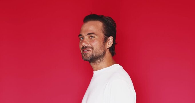 Handsome bearded young man with stylish slicked back dark hair wearing white t shirt turning head and winking with playful mischievous facial expression and charming smile isolated on red background