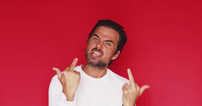 Handsome bearded man wearing white t shirt looking at camera, smiling with happy joyful facial expression after showing middle finger gesture with aggressive and angry mood isolated on red background