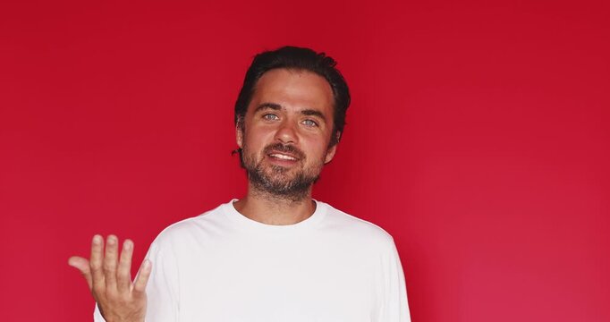 Angry bearded man wearing white t shirt looking at camera and arguing aggressively with furious facial expression and emotional shouting gesturing with hands isolated on a solid red studio background 