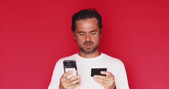 Handsome bearded man calm and neutral facial expression after looking at smartphone and credit card with disappointed and frustrated mood isolated on a solid red studio background close up portrait