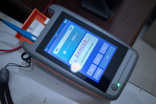 An optical time-domain reflectometer testing fiber optic cables, displaying a laser danger warning on screen.