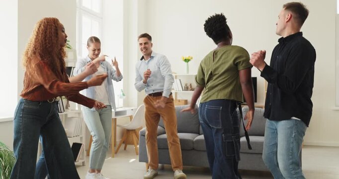 Young friends dancing at home party, fun celebration. Diverse group of people moves to music in a living room, happy and clapping together. Joyful home celebration and friendship concept.