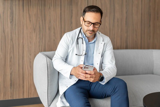 Doctor sitting on a couch, holding a smartphone, representing modern healthcare, digital communication, and telehealth services in a clinic or office setting