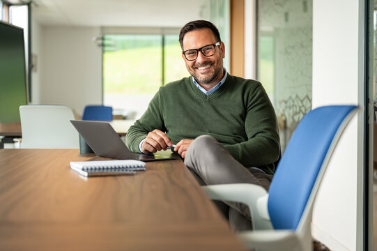 Happy male professional smiling at the camera, holding a stylus pen while working with technology at a desk, embodying confidence, success, and focused productivity in a contemporary workspace