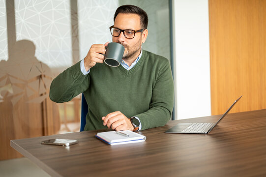 Man taking a moment to drink coffee while working at a conference table, utilizing technology for productivity in a contemporary business setting