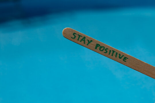 Woman holding a popsicle stick with the words "Stay positive" against a blue swimming pool background, creating an uplifting summer lifestyle concept about optimism, happiness and positive thinking.