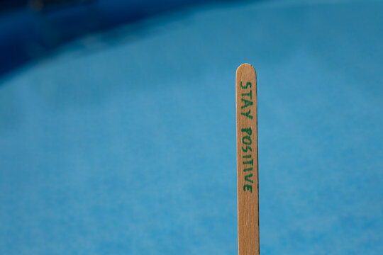 Woman holding a popsicle stick with the words "Stay positive" against a blue swimming pool background, creating an uplifting summer lifestyle concept about optimism, happiness and positive thinking.