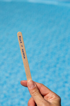 Woman holding a popsicle stick with the words "Make it happen" against a blue swimming pool background, creating a motivational concept about taking action, achieving goals and turning ideas into real