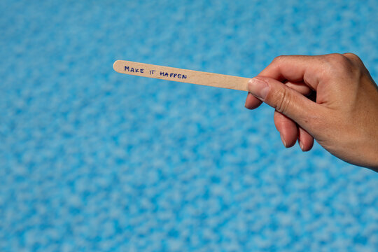 Woman holding a popsicle stick with the words "Make it happen" against a blue swimming pool background, creating a motivational concept about taking action, achieving goals and turning ideas into real