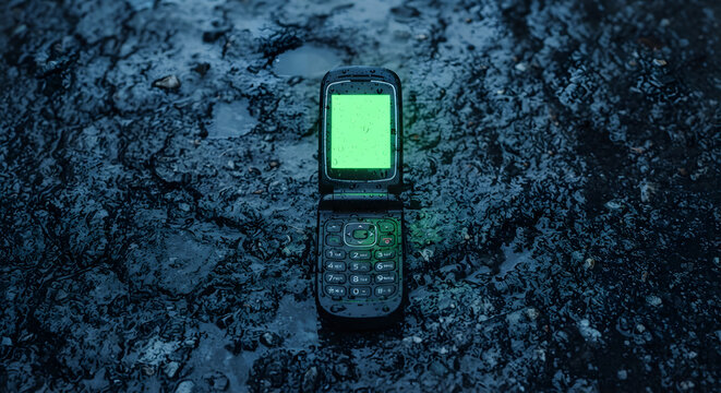 Abandoned flip phone with green screen on wet asphalt
