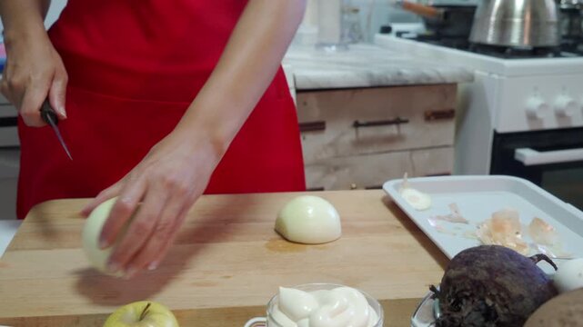 A woman in a red apron carefully slices an onion on a wooden board. She works with calm focus in a clean, bright home kitchen.