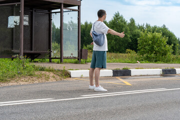 Man with a bag hitchhikes on a rural road by extending his thumb. The wide road stretches into the distance with a vehicle approaching, highlighting travel and adventure themes. © magpie