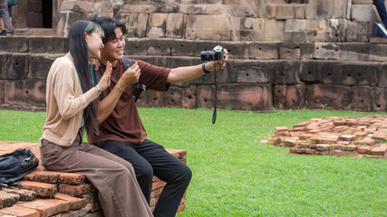 Fototapeta premium Young Asian couple taking selfie video with camera mounted on small tripod, sitting on ancient stone ruins terrace, enjoying fun romantic sightseeing vacation at heritage park.