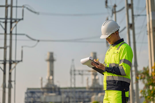 Asian male electrical engineer holding digital tablet standing at power plant station. Professional energy industry technician working on infrastructure maintenance and renewable resource project.