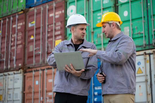 Two professional male logistics engineers use a digital laptop while standing in a busy commercial shipping container yard discussing cargo transport operations for a global supply chain.