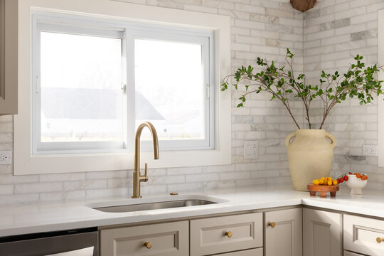 A gold kitchen faucet detail in a luxury kitchen with brown cabinets, marble subway tile backsplash, and decorations on the marble countertop.