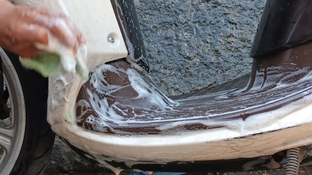 A scooter footboard covered with cleaning foam during a vehicle wash. Automotive maintenance concept with detailed soap texture and outdoor setting.