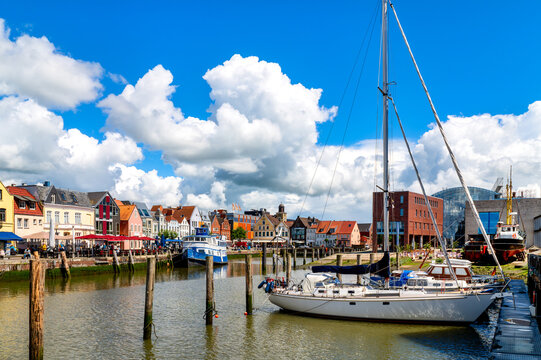 Sailing yacht in the harbor of Husum with the historic old town, waterfront promenade and dramatic clouds beneath a blue sky.