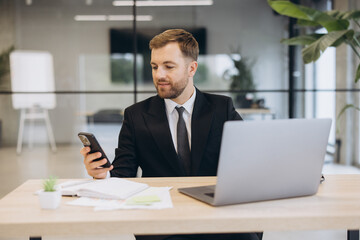Obraz premium Businessman managing communication on smartphone while working on laptop in modern office environment