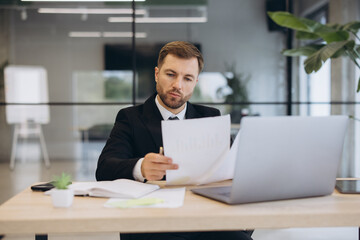 Fototapeta premium Focused businessman analyzing financial documents and data on laptop in modern office, managing business growth