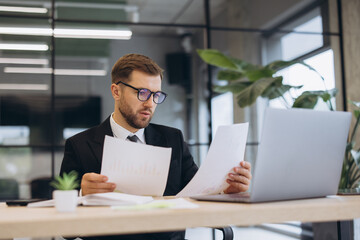 Young businessman reviewing financial reports in office, analyzing documents and working on laptop