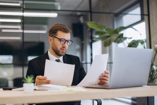 Young businessman reviewing financial reports in office, analyzing documents and working on laptop