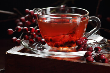 Healthy hawthorn berry tea in a glass cup surrounded by red fruit on a dark moody background © spiegelka