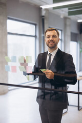 Fototapeta premium Businessman smiling, attaching sticky notes to a glass wall, planning project strategy in modern office