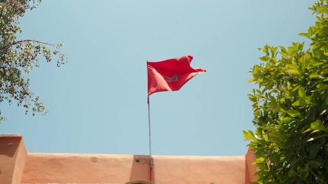Red Flag Waving Over Terracotta Rooftop Against Blue Sky With Green Foliage