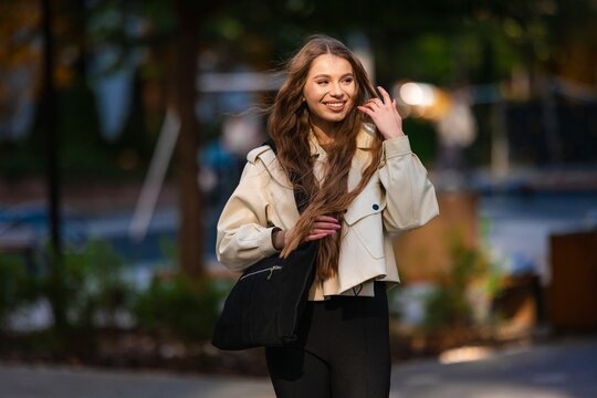 Smiling woman wearing beige trench coat outdoors park sunlight portrait warm cheerful expression natural styling fashion accessory shoulder bag casual urban setting