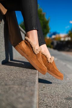 Brown suede shearling moccasin boot beside leg in black pants on stone steps under bright blue sky, close view showing stitching, cozy lining, urban casual style and soft winter comfort